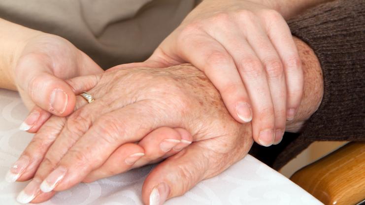 Zwei Hände, eine ältere und eine jüngere, liegen auf einem Tisch, wobei die jüngere Hand die ältere sanft umschließt.