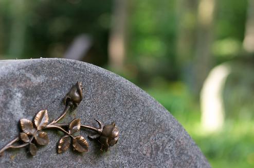 Bronze-Rosenornament auf einer grauen Grabsteinkante, unscharfer Hintergrund mit grünem Waldboden.