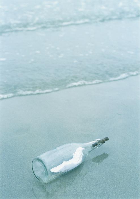 Eine Glasflasche liegt am Strand, umgeben von sanften Wellen und feinem Sand. In der Flasche ist ein dünnes Papier.
