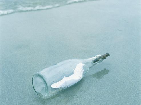 Eine Glasflasche liegt am Strand, umgeben von sanften Wellen und feinem Sand. In der Flasche ist ein dünnes Papier.