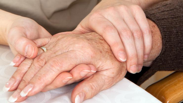 Zwei Hände, eine ältere und eine jüngere, liegen auf einem Tisch, wobei die jüngere Hand die ältere sanft umschließt.